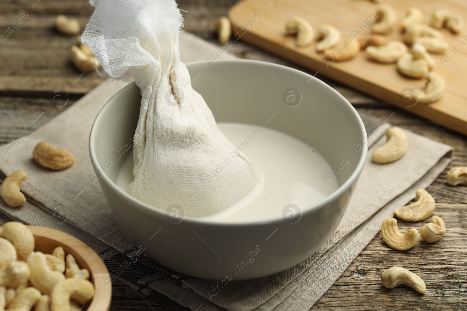 Photo of Making cashew milk. Cheesecloth and milk in bowl among nuts on wooden table, closeup