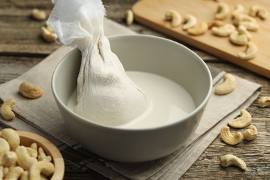 Photo of Making cashew milk. Cheesecloth and milk in bowl among nuts on wooden table, closeup