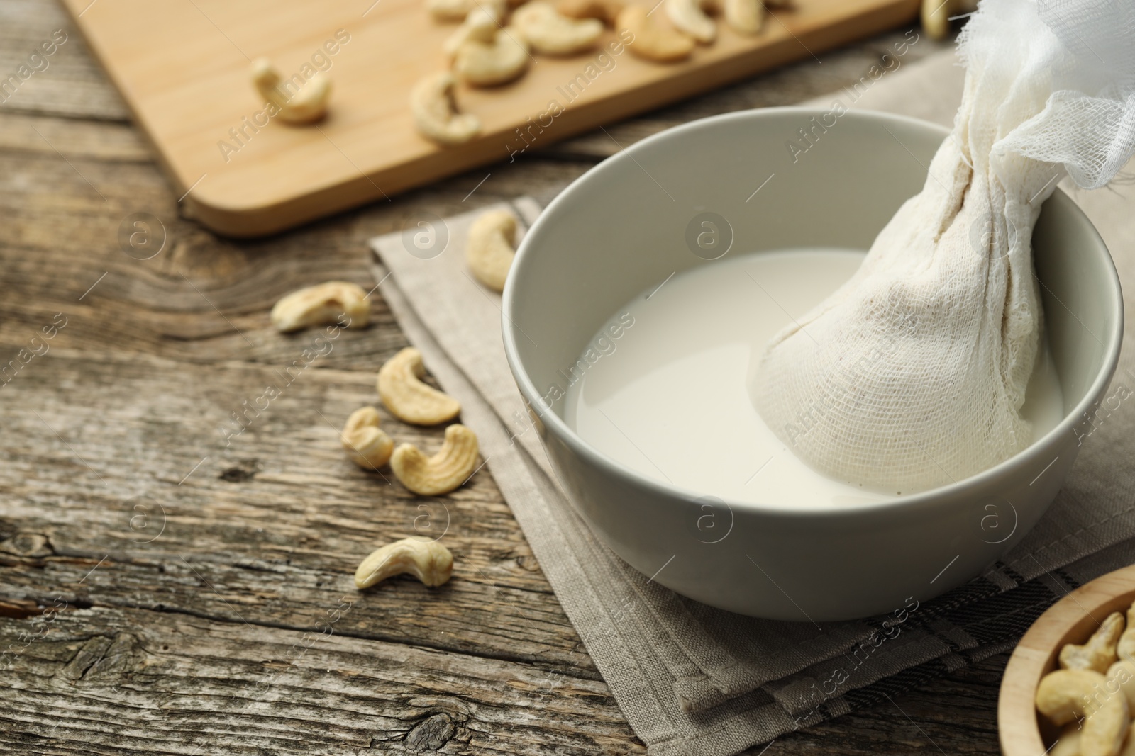 Photo of Making cashew milk. Cheesecloth and milk in bowl among nuts on wooden table, closeup