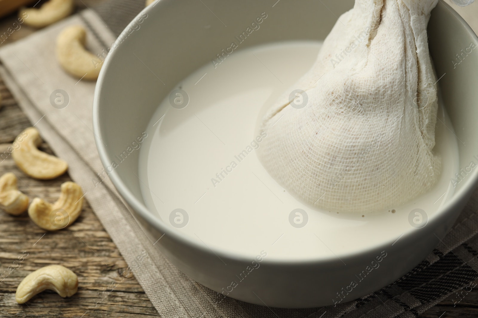 Photo of Making cashew milk. Cheesecloth and milk in bowl among nuts on wooden table, closeup