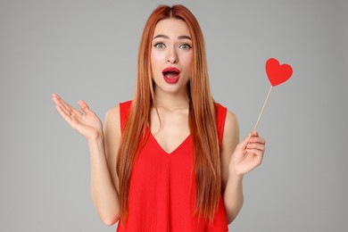 Photo of Emotional young woman in red dress with paper heart on light grey background