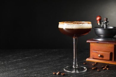 Photo of Glass of delicious espresso martini, manual grinder and coffee beans on dark textured table, closeup. Space for text