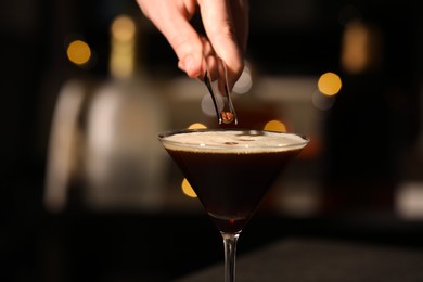 Photo of Bartender putting coffee bean into glass with delicious espresso martini against blurred lights, closeup