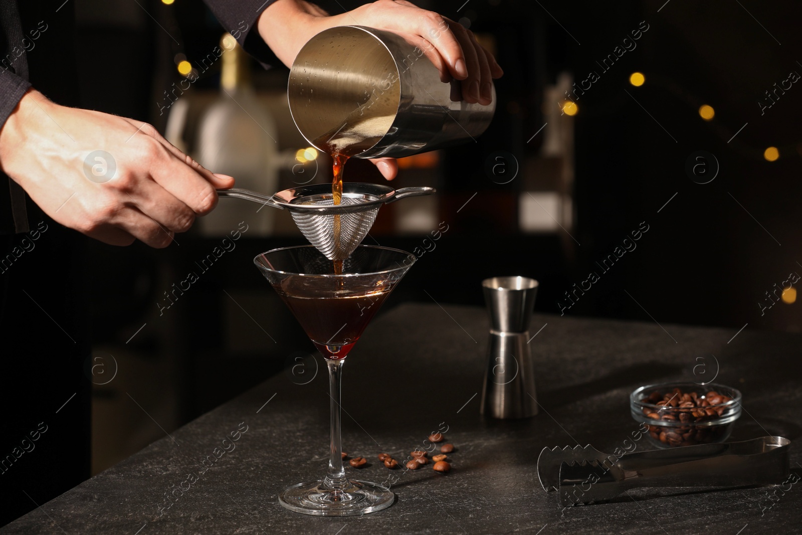 Photo of Bartender making delicious espresso martini at dark table against blurred lights, closeup