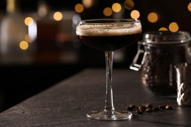 Photo of Glass with delicious espresso martini and coffee beans on dark table against blurred lights, closeup. Space for text