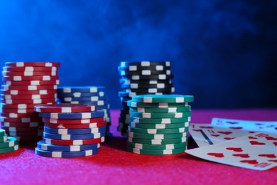 Photo of Poker chips and playing cards on pink table in neon lights, closeup