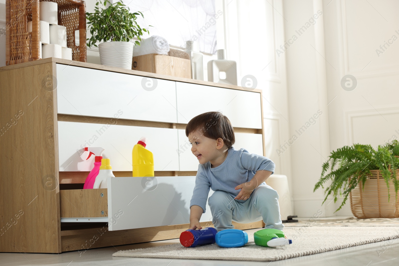 Photo of Little boy near cabinet with bottles of detergents at home. Child in danger