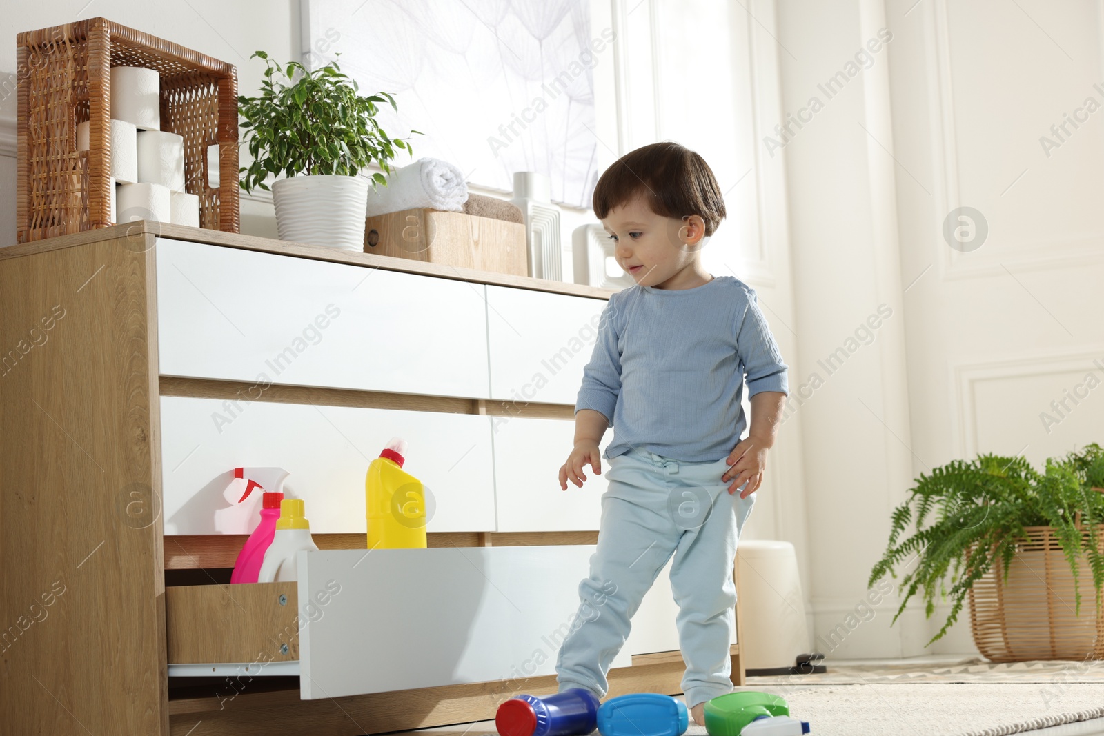 Photo of Little boy near cabinet with bottles of detergents at home. Child in danger