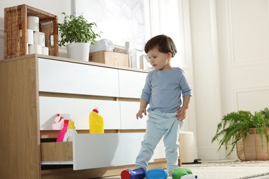 Photo of Little boy near cabinet with bottles of detergents at home. Child in danger