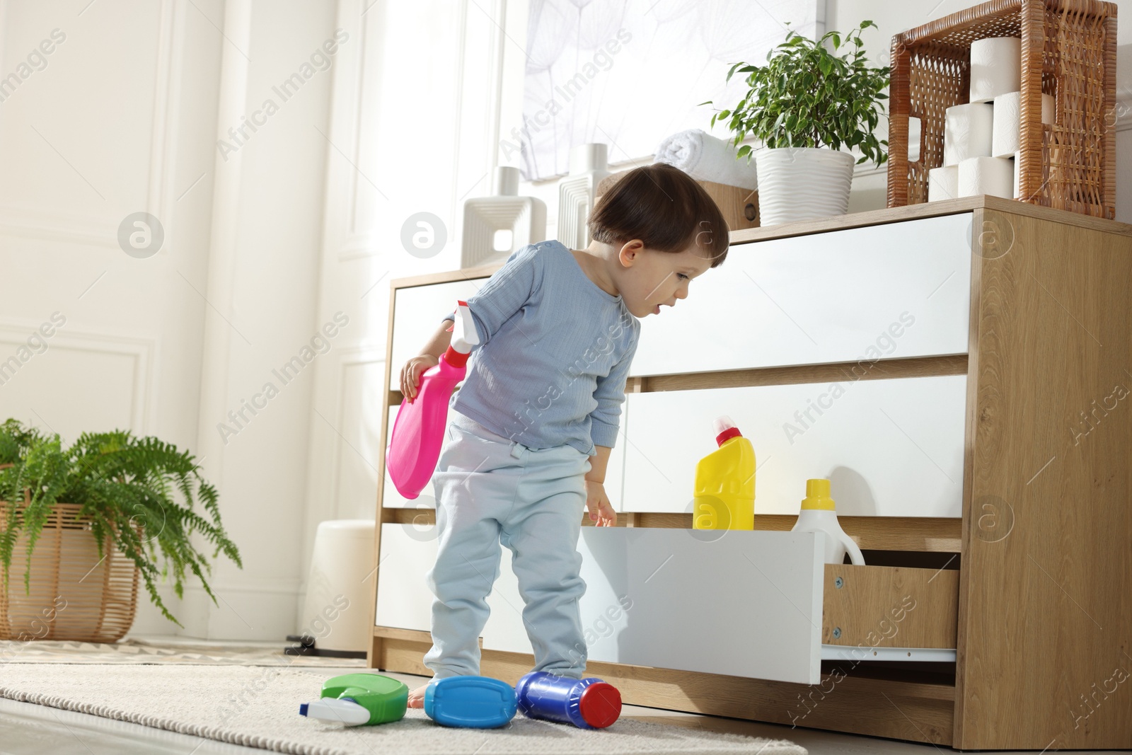 Photo of Little boy opening cabinet with bottles of detergents at home. Child in danger