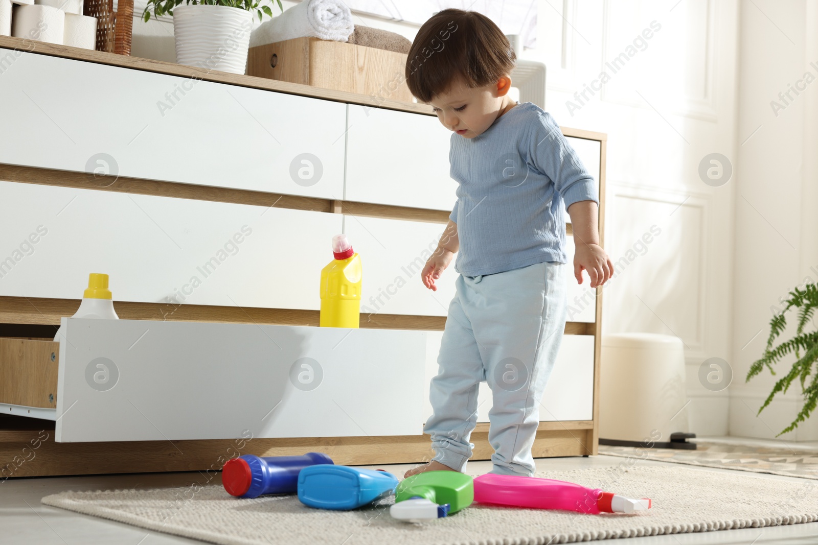 Photo of Little boy playing with bottles of detergents near cabinet at home. Child in danger