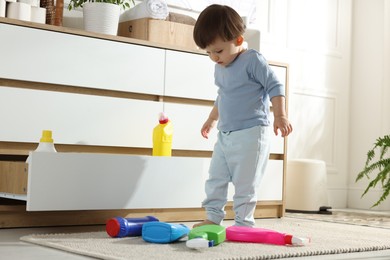 Photo of Little boy playing with bottles of detergents near cabinet at home. Child in danger