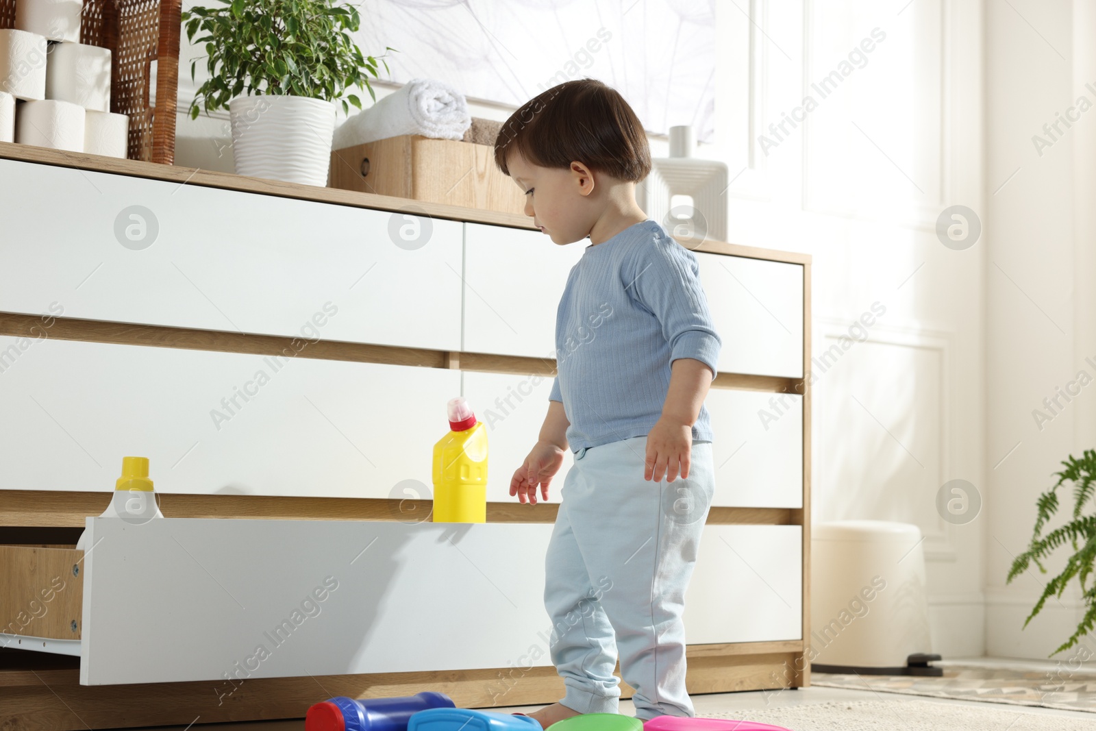 Photo of Little boy near cabinet with bottles of detergents at home. Child in danger