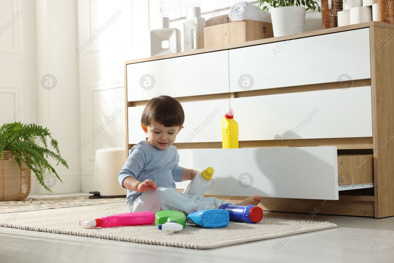 Photo of Little boy playing with bottles of detergents near cabinet at home. Child in danger