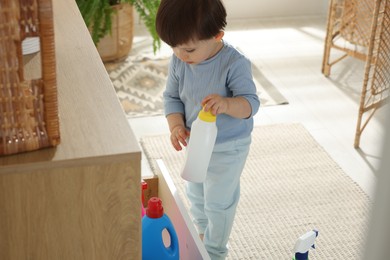 Photo of Little boy playing with bottles of detergents near cabinet at home. Child in danger