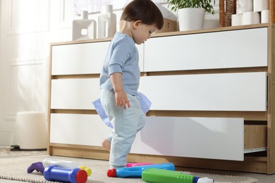 Photo of Little boy playing with bottles of detergents near cabinet at home. Child in danger