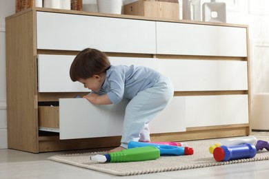 Photo of Little boy opening drawer with bottles of detergents at home. Child in danger