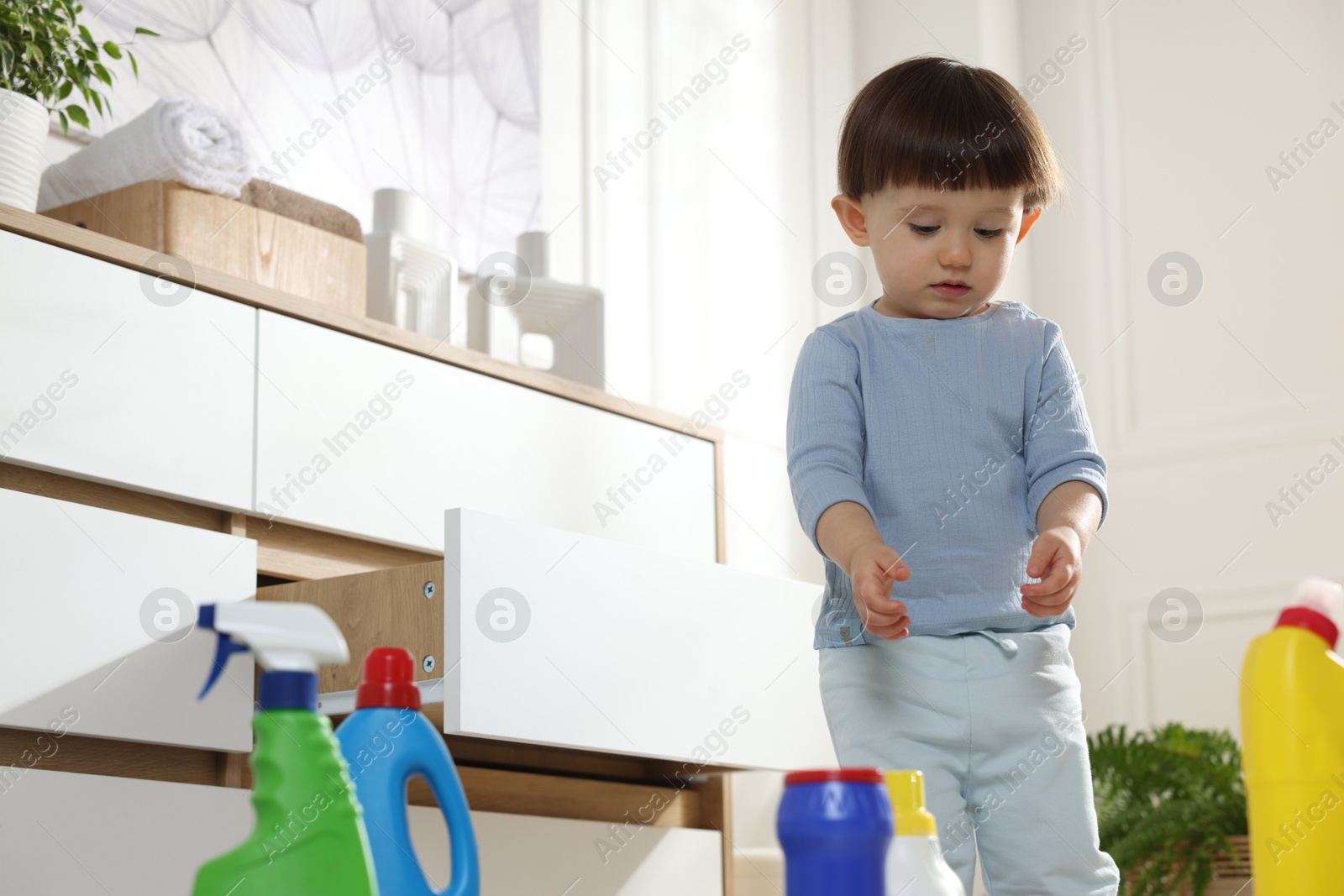 Photo of Little boy playing with bottles of detergents near cabinet at home. Child in danger