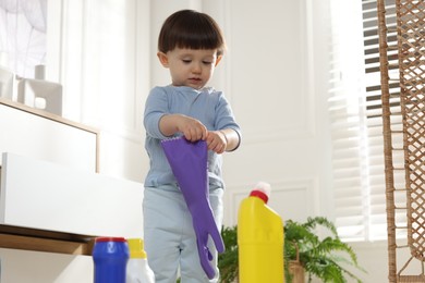 Photo of Little boy playing with glove among bottles of detergents near cabinet at home. Child in danger