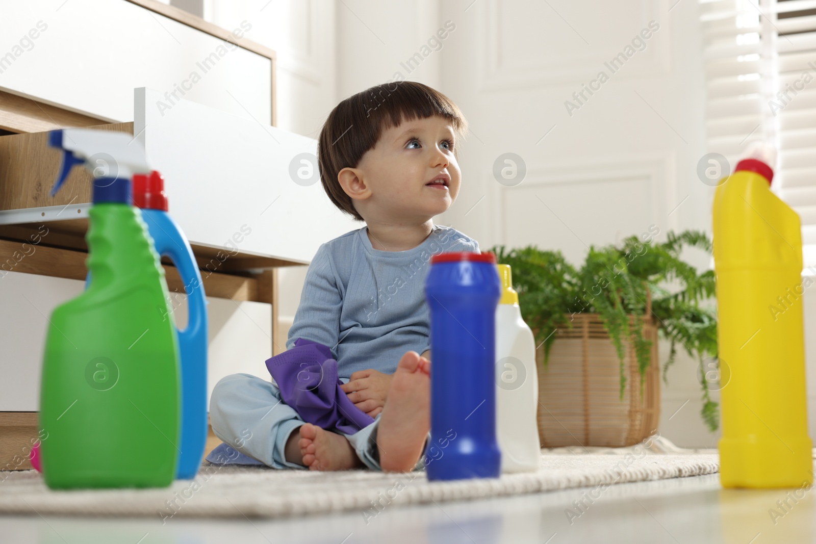 Photo of Little boy playing with glove among bottles of detergents near cabinet at home. Child in danger