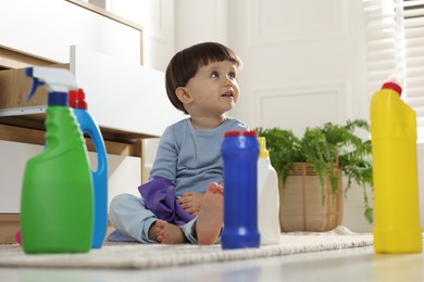 Photo of Little boy playing with glove among bottles of detergents near cabinet at home. Child in danger