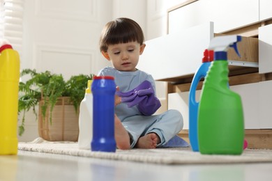 Photo of Little boy playing with glove among bottles of detergents near cabinet at home. Child in danger