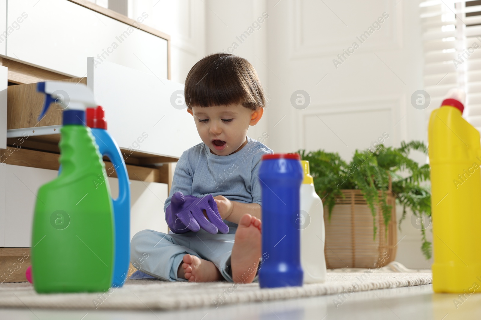 Photo of Little boy playing with glove among bottles of detergents near cabinet at home. Child in danger