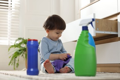 Photo of Little boy playing with glove among bottles of detergents near cabinet at home. Child in danger