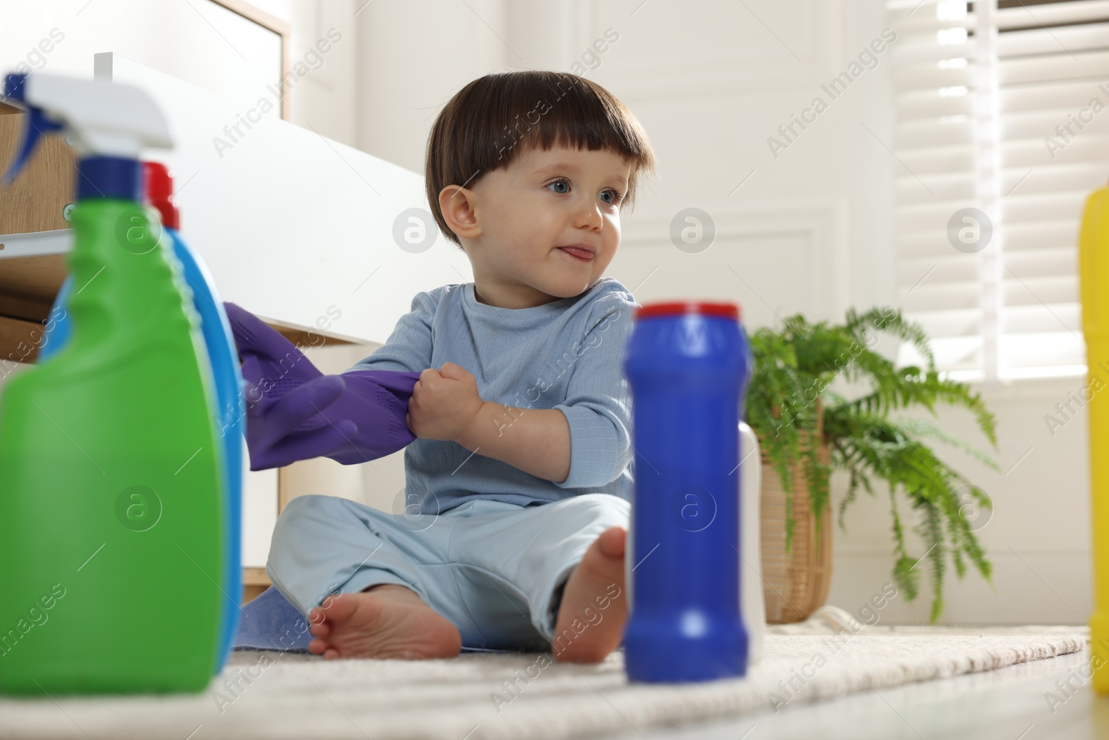 Photo of Little boy playing with glove among bottles of detergents near cabinet at home. Child in danger