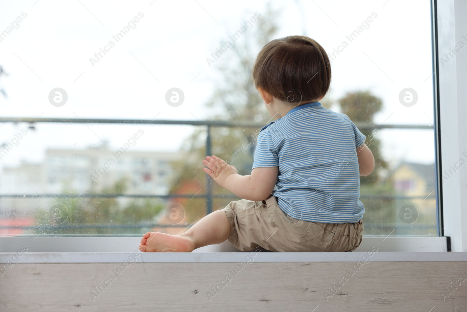 Photo of Little boy sitting on window sill, back view. Child in danger