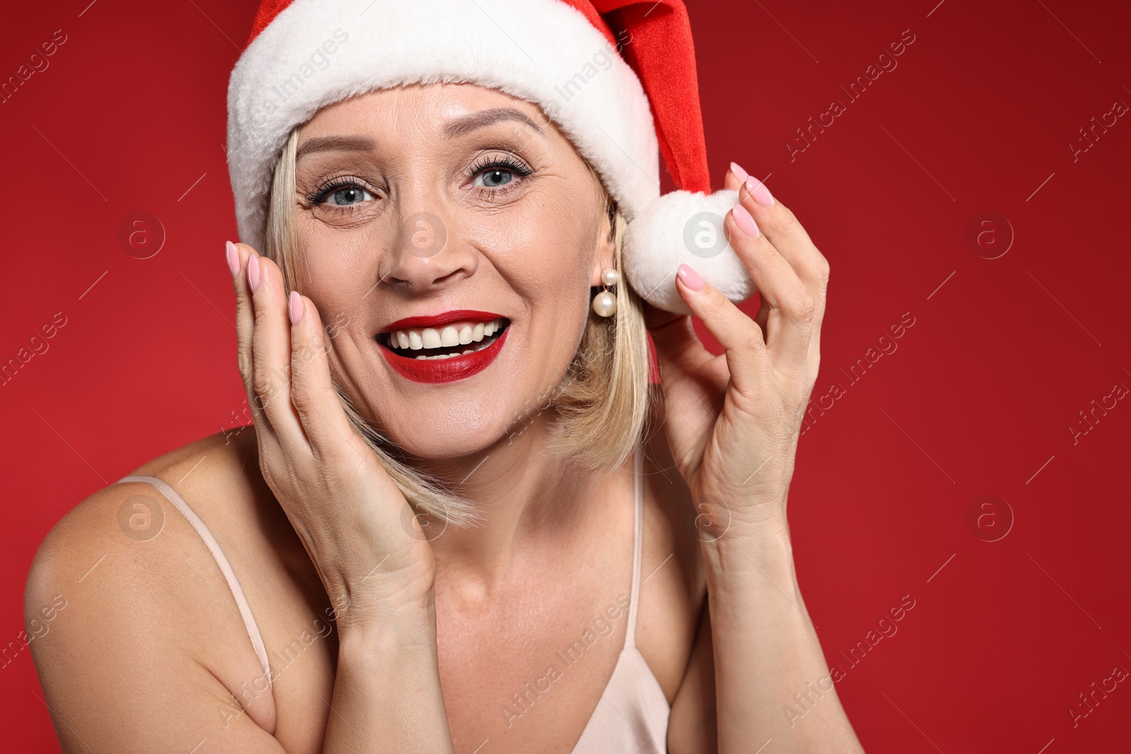 Photo of Smiling woman with perfect makeup in Santa hat celebrating Christmas on red background