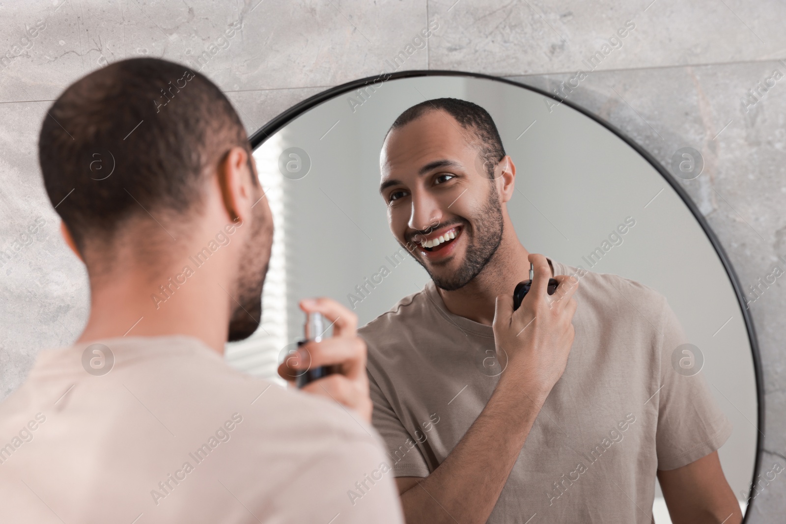 Photo of Smiling man spraying perfume near mirror in bathroom