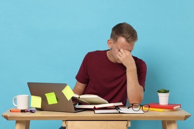 Photo of Preparing for exam. Tired student with laptop and books at table against light blue background