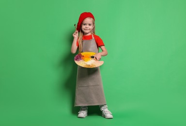 Photo of Little girl with brush and palette pretending to be artist on green background. Dreaming of future profession