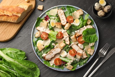 Photo of Tasty Caesar salad with chicken and cutlery on dark gray textured table, flat lay