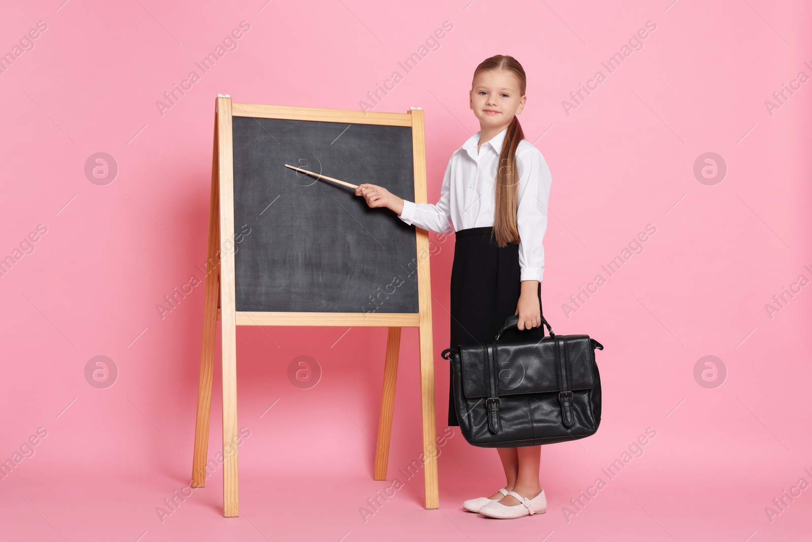 Photo of Little girl with pointer and briefcase near chalkboard pretending to be teacher on pink background. Dreaming about future profession