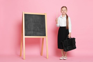 Photo of Little girl with pointer and briefcase near chalkboard pretending to be teacher on pink background. Dreaming about future profession