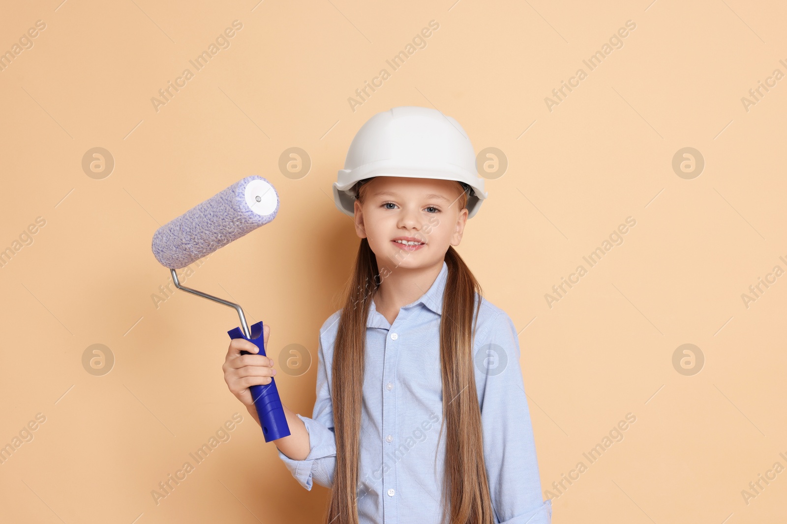 Photo of Little girl in hard hat with roller brush on beige background. Dreaming about future profession