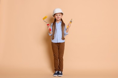 Photo of Little girl in hard hat with painting tools on beige background. Dreaming about future profession