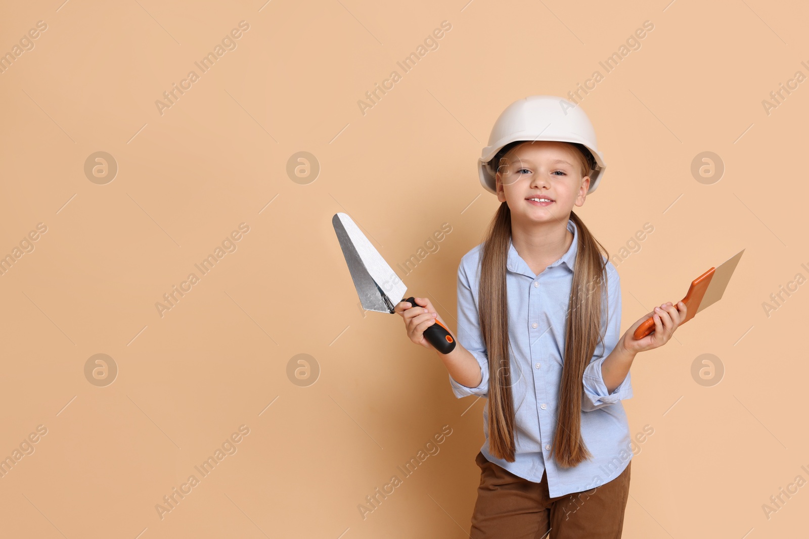 Photo of Little girl in hard hat with putty knife and trowel on beige background, space for text. Dreaming about future profession