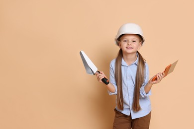 Photo of Little girl in hard hat with putty knife and trowel on beige background, space for text. Dreaming about future profession