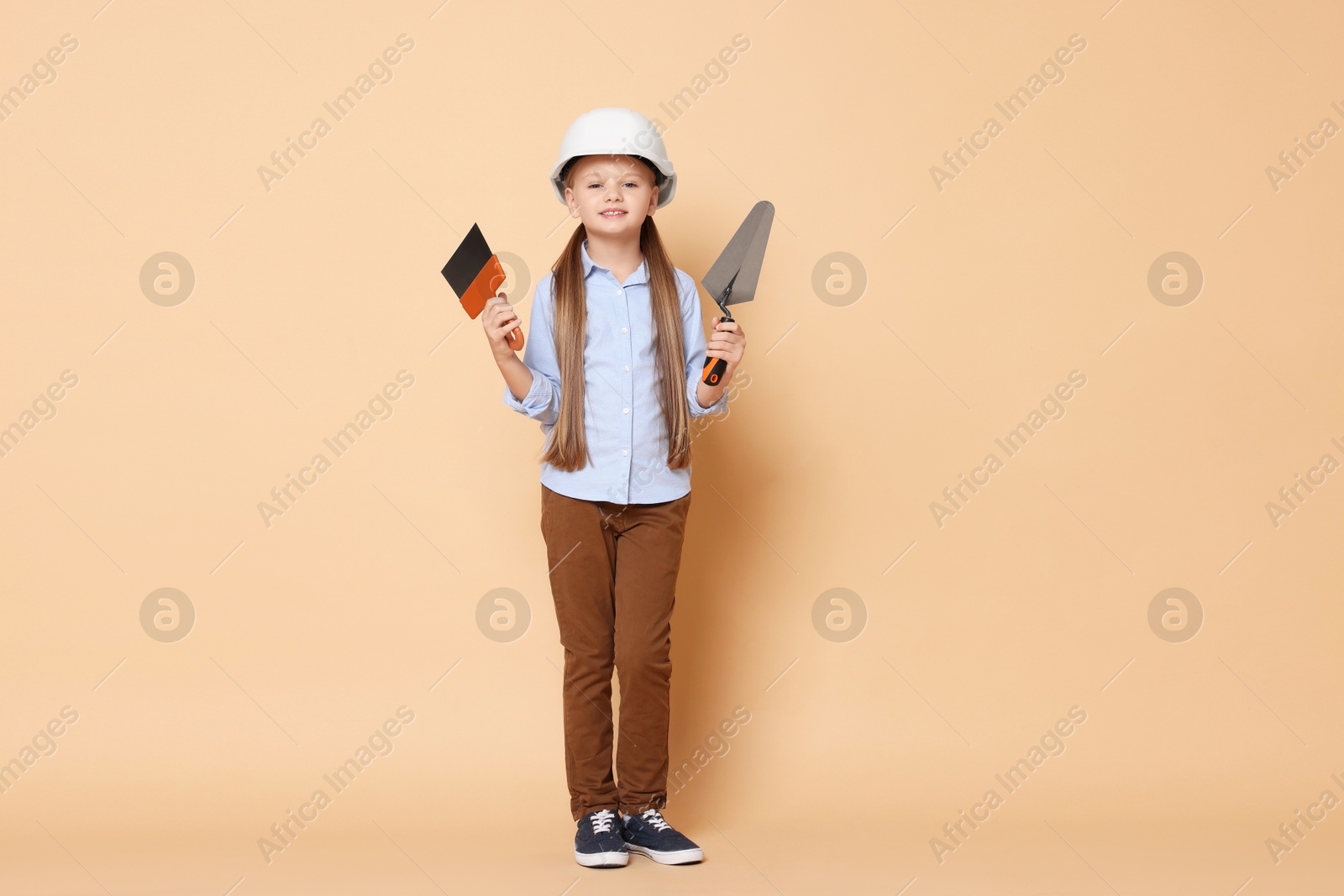 Photo of Little girl in hard hat with putty knife and trowel on beige background. Dreaming about future profession