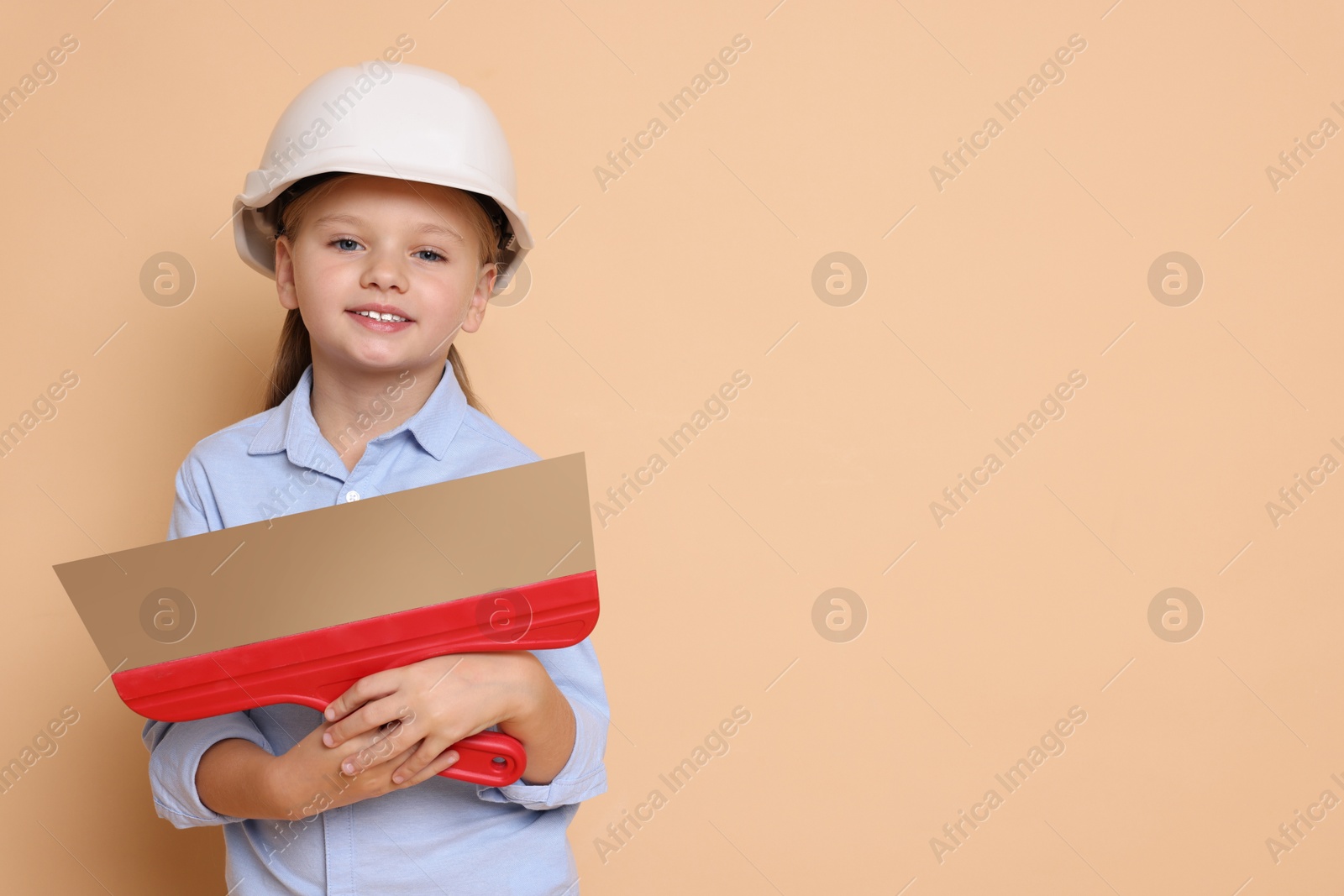 Photo of Little girl in hard hat with putty knife on beige background, space for text. Dreaming about future profession