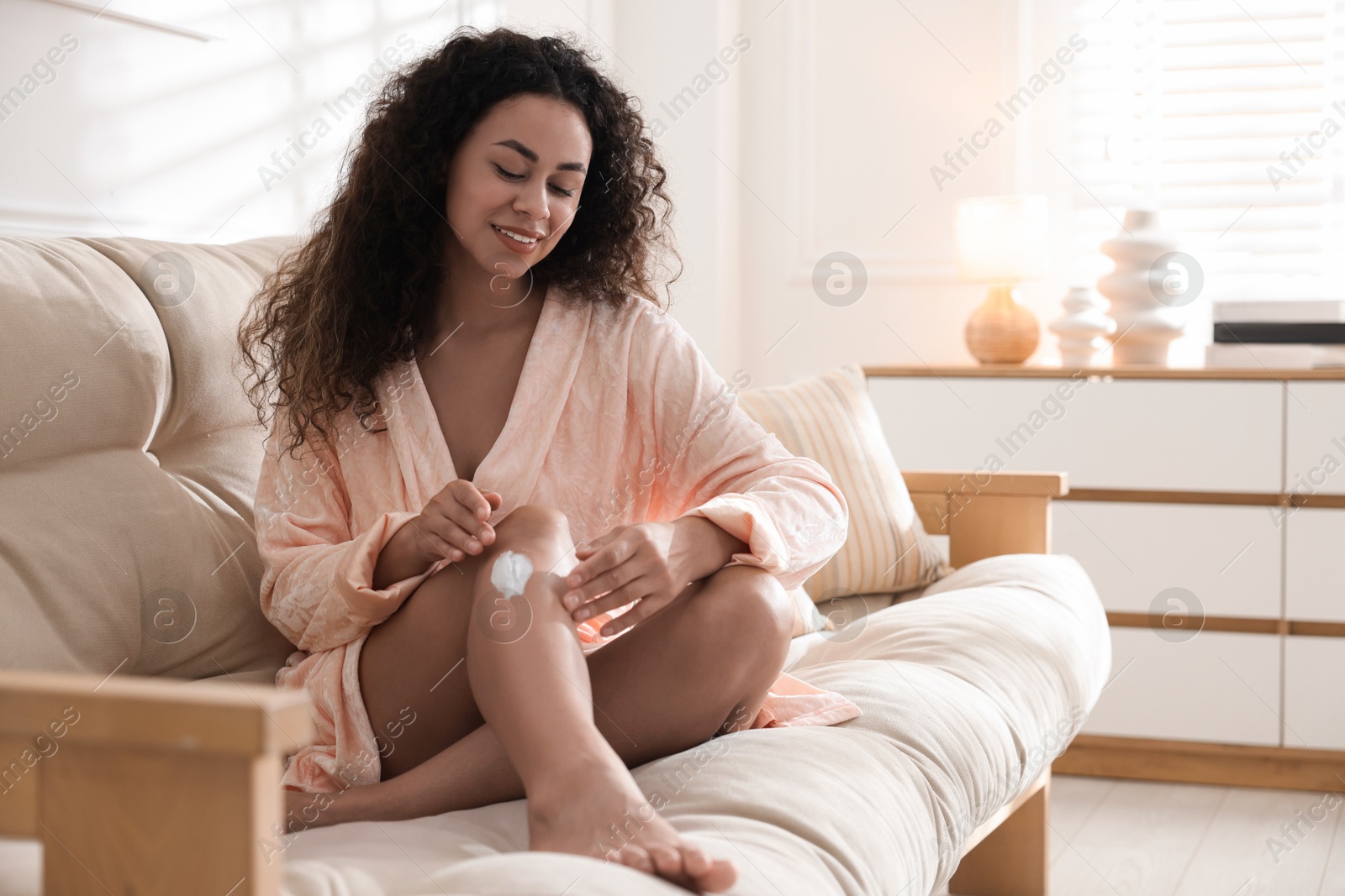 Photo of Young woman applying cream onto leg on sofa at home. Space for text