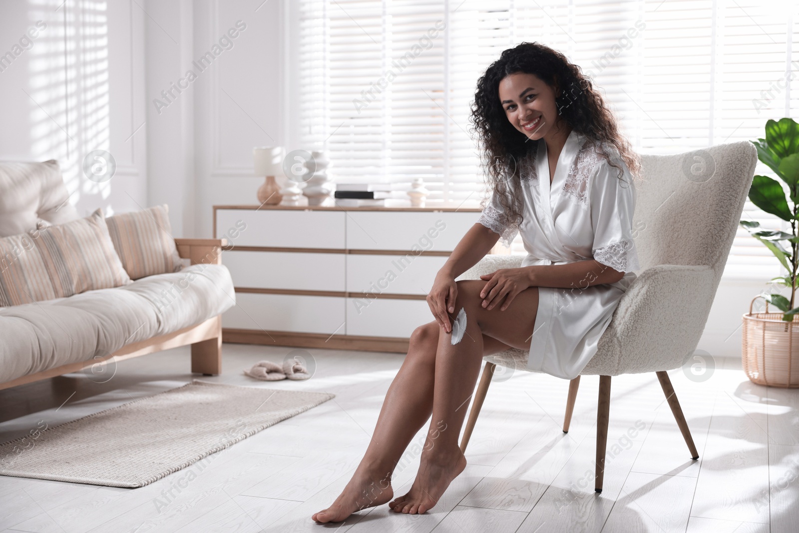 Photo of Young woman applying cream onto leg in armchair at home