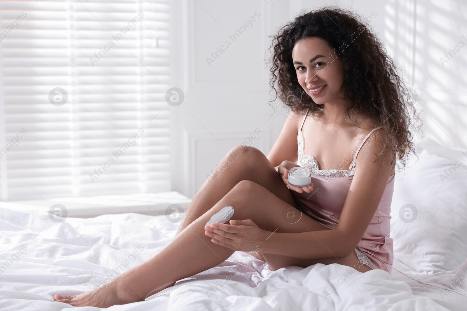 Photo of Young woman applying cream onto leg on bed at home. Space for text