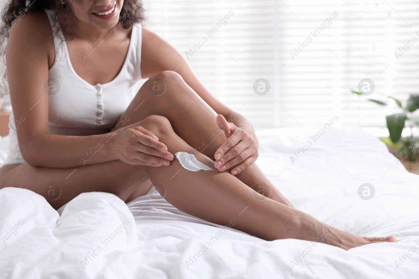 Photo of Woman applying cream onto leg on bed indoors, closeup