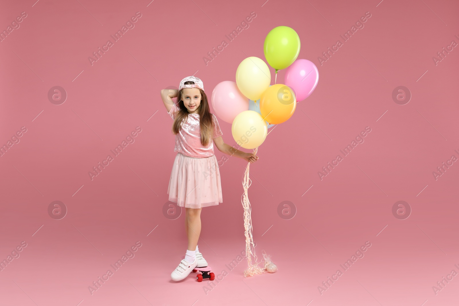 Photo of Little girl with colorful balloons and penny board on pink background