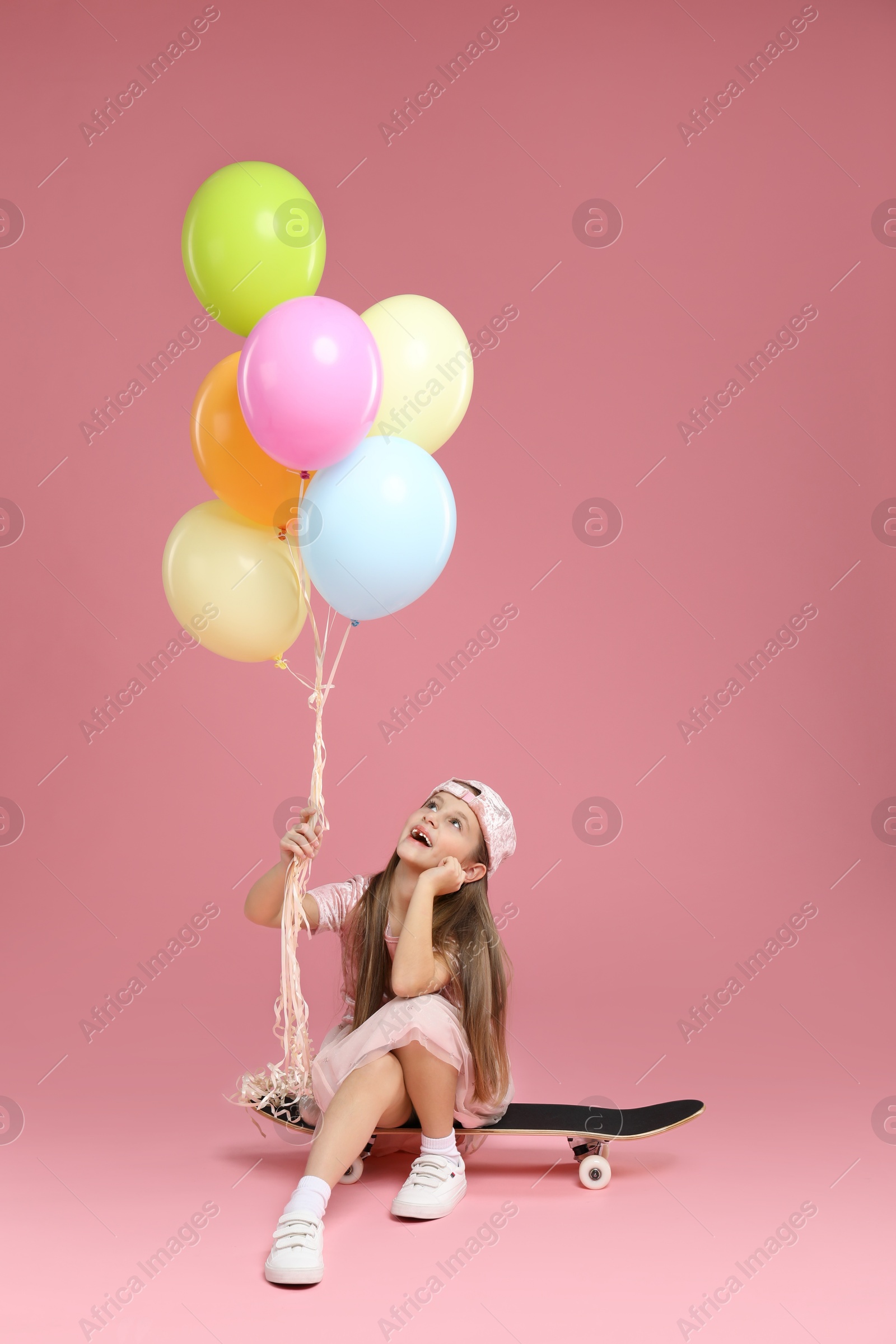 Photo of Little girl with colorful balloons and skateboard on pink background