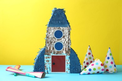 Photo of Beautiful pinata in shape of rocket, party cones and poppers on blue table against yellow background